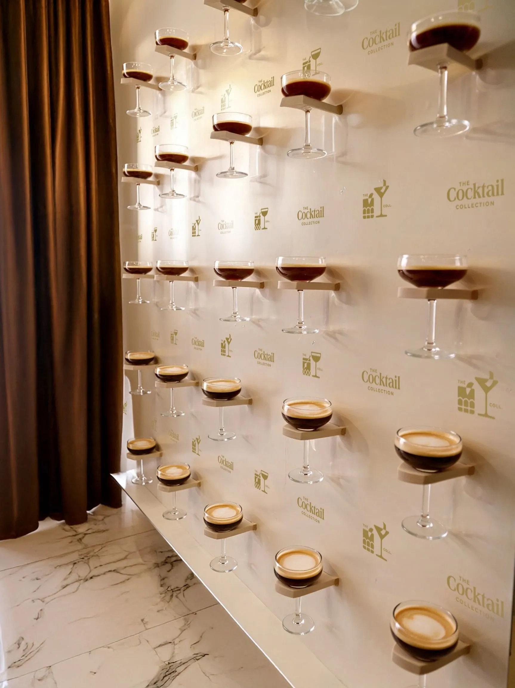 The espresso martini wall: coupes on shelves against a wall patterned with The Cocktail Collection wordmark and icons