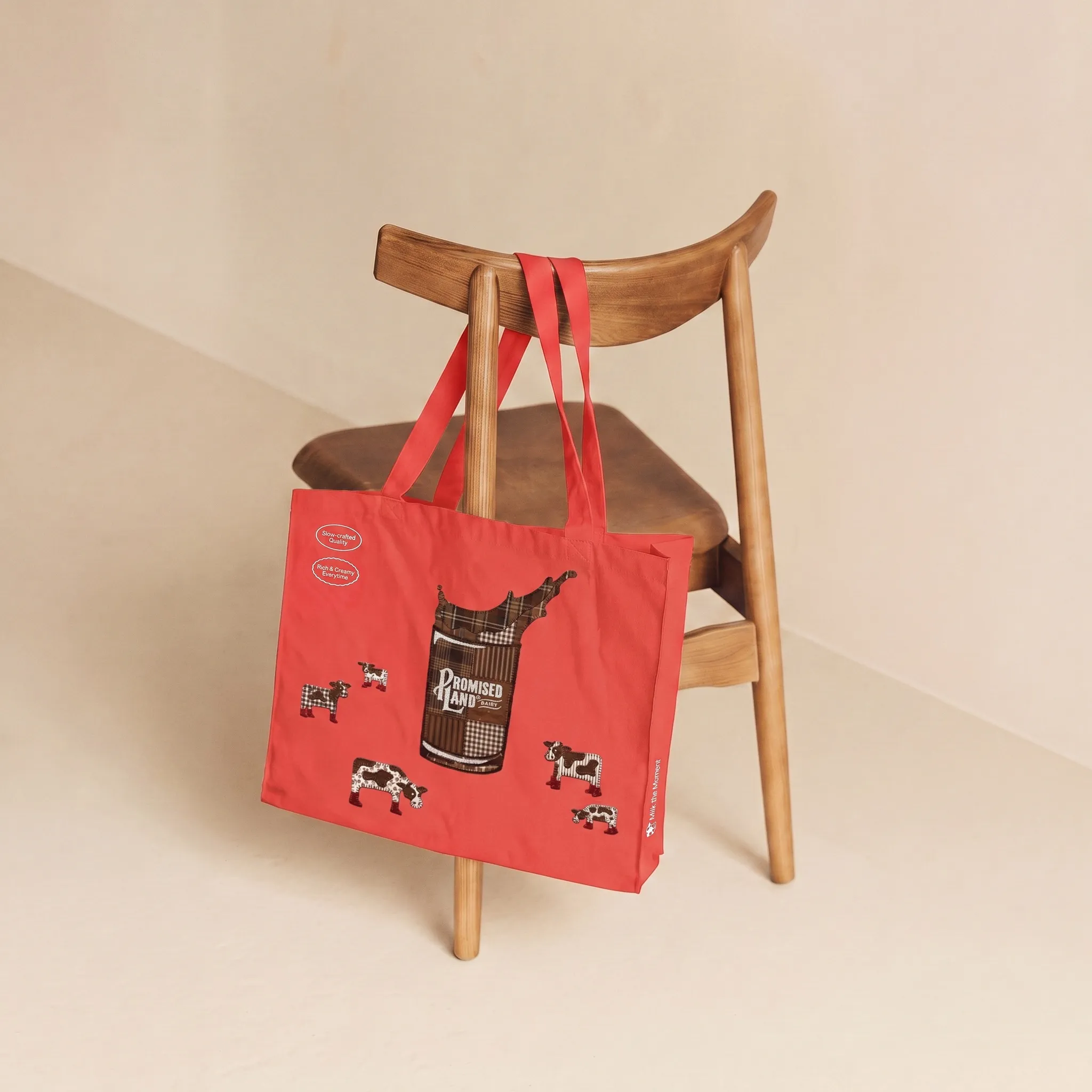 Red Promised Land Dairy tote bag hanging on a wooden chair: a patchwork glass of milk and cows in red boots, “Slow-crafted Quality”, “Rich & Creamy Everytime”, “Milk the Moment”