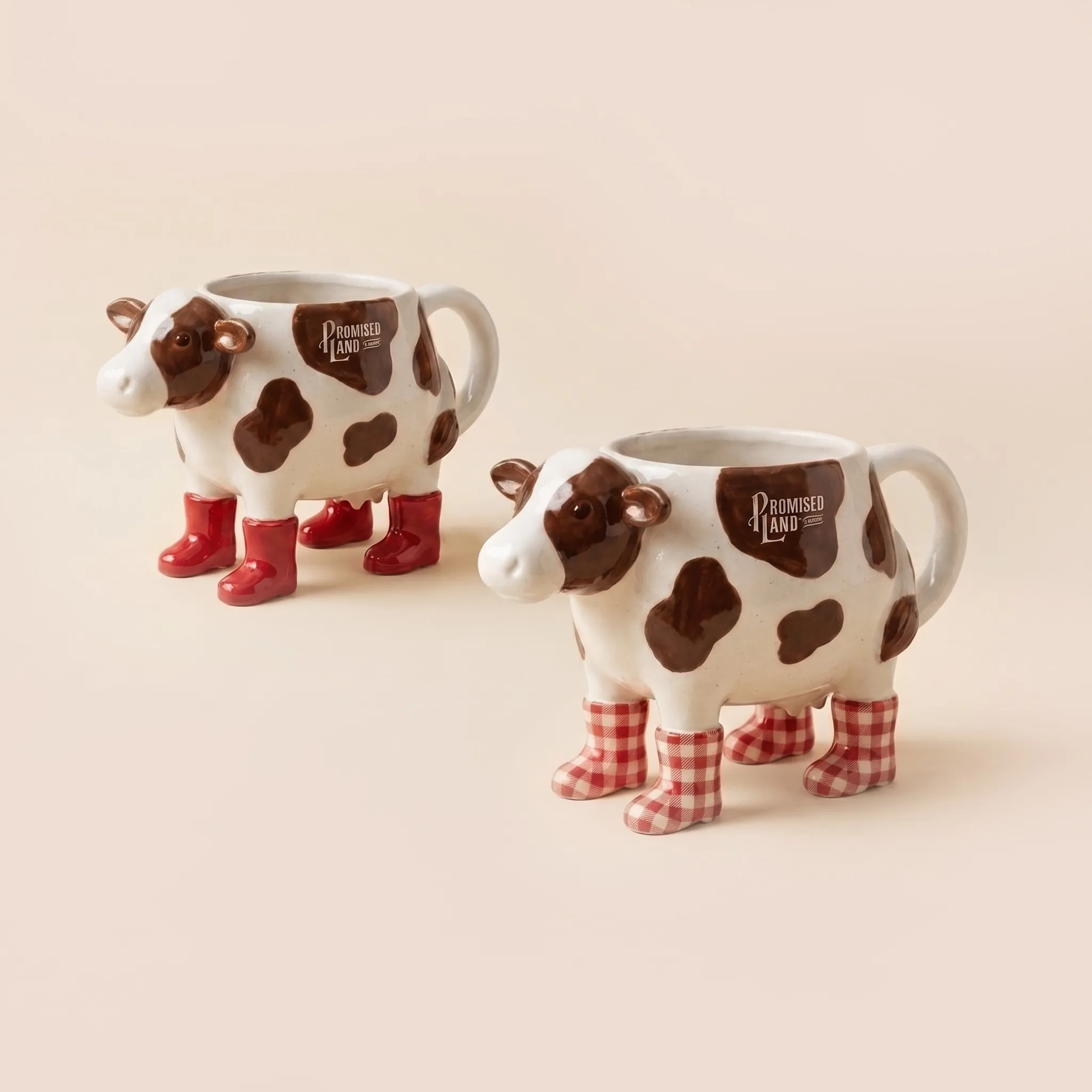 Two cow-shaped ceramic mugs with the Promised Land Dairy logo, one in red boots and one in red gingham boots