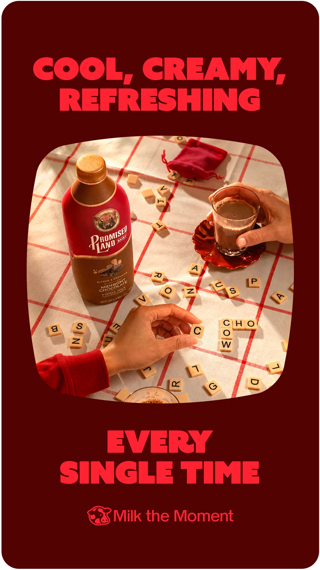 Social post on dark red reading “Cool, creamy, refreshing” and “Every single time”, over a photo of two people at a word-tile game on a red-striped cloth with a bottle of Promised Land Dairy Midnight Chocolate whole milk and a glass of chocolate milk, the tiles spelling “COW” and “CHO”; signed “Milk the Moment”